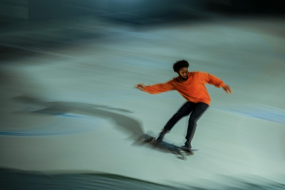 High angle of African American man in casual clothes stretching out arms and riding longboard fast in evening in skatepark.