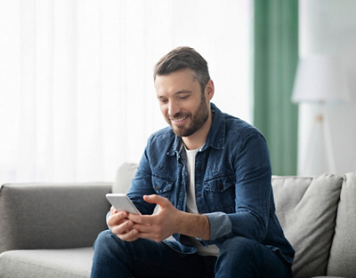 Mann sitzt auf dem Sofa und bedient lächelnd seit Mobiltelefon