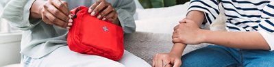 Parent opening a small red first aid kit while comforting a child at home / Group of hikers walking through dense forest with a red first aid kit attached to a backpack / Person taking a red first aid kit out of a car’s glove compartment