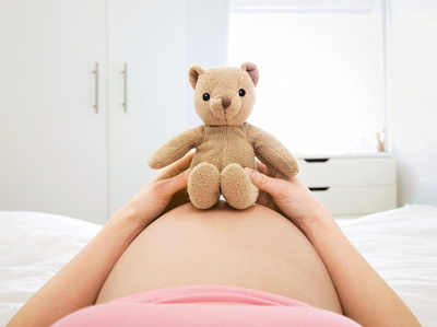 Pregnant woman laying on bed with teddy bear on belly, mid section