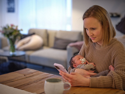 Eine Frau sitzt an einem Tisch am Smartphone und hält ihr Baby auf dem Arm..
