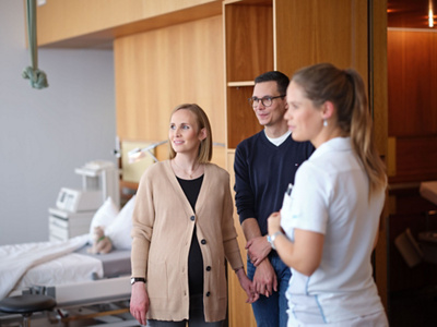 Eine schwangere Frau besichtigt mit Ihrem Mann den Gebärsaal der Klinik St. Anna und sie werden von einer Fachperson begleitet.