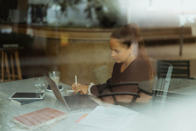Fabienne sitzt konzentriert in einem Kaffee und macht Notizen, während sie auf ihren Laptop blickt.