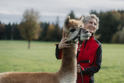 Miriam steht mit ihrem Lama auf einer grünen Wiese und lacht fröhlich.